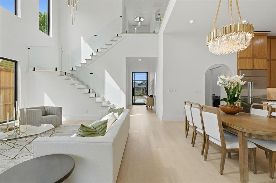 Expansive open-concept living area featuring a dramatic floating staircase with glass railings, double-height ceilings, large windows, light wood-finish flooring, and a contemporary chandelier Expansive open-concept living area featuring a dramatic floating staircase with glass railings, double-height ceilings, large windows, light wood-finish flooring, and a contemporary chandelier