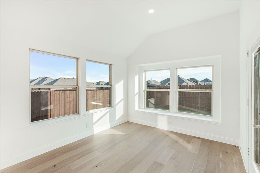 Empty room with vaulted ceiling, light wood floors, and multiple windows overlooking a fenced backyard
