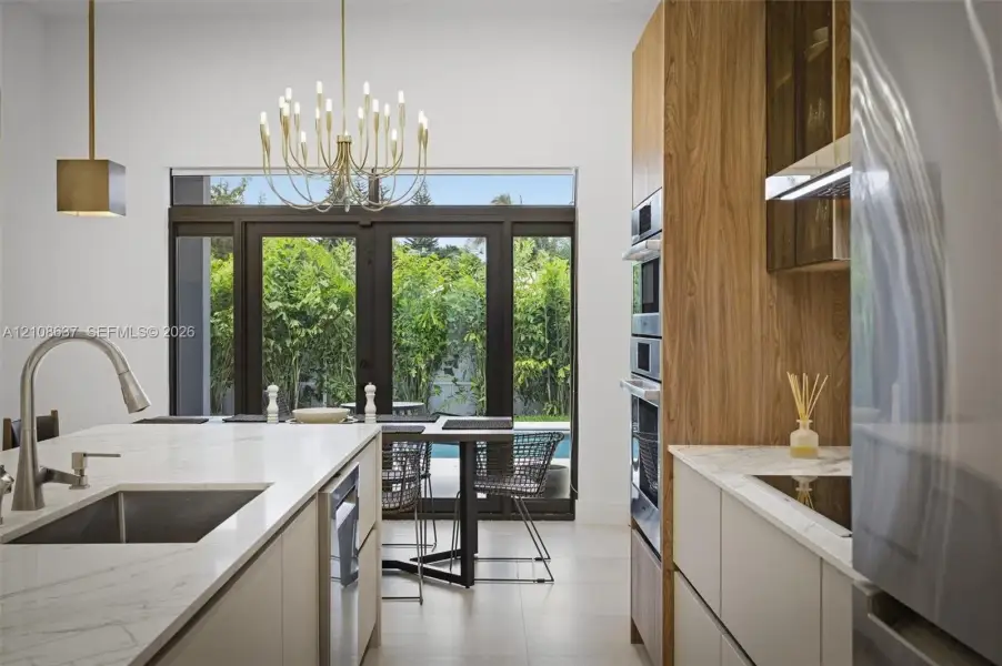 Kitchen featuring a central island with a stone-finish countertop and undermount sink, flat-panel cabinetry with wood-grain accents, built-in stainless steel double ovens, and large-format tile flooring