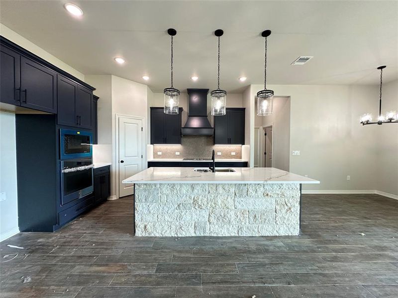 Kitchen featuring light stone counters, wall chimney range hood, oven, decorative light fixtures, and a kitchen island with sink Kitchen featuring light stone counters, wall chimney range hood, oven, decorative light fixtures, and a kitchen island with sink