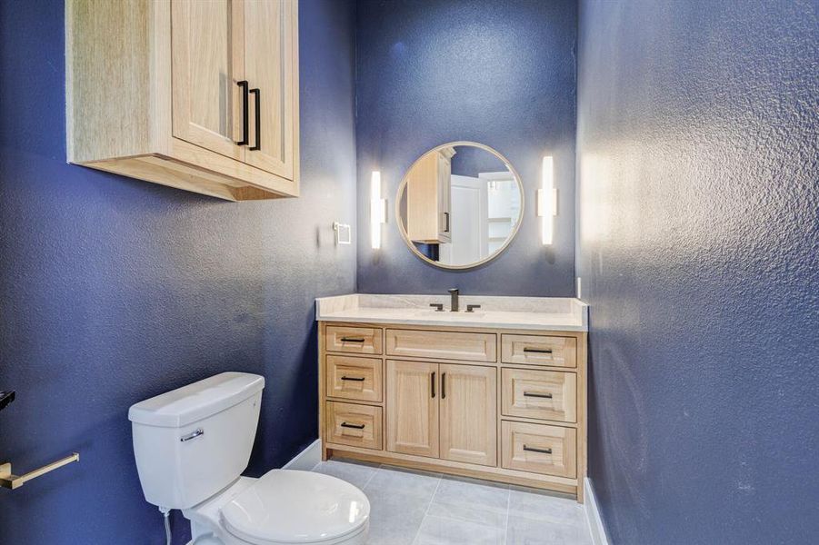 Bathroom vanity featuring light wood-finish cabinetry with black hardware, a light-toned countertop, and a round mirror with vertical sconce lighting