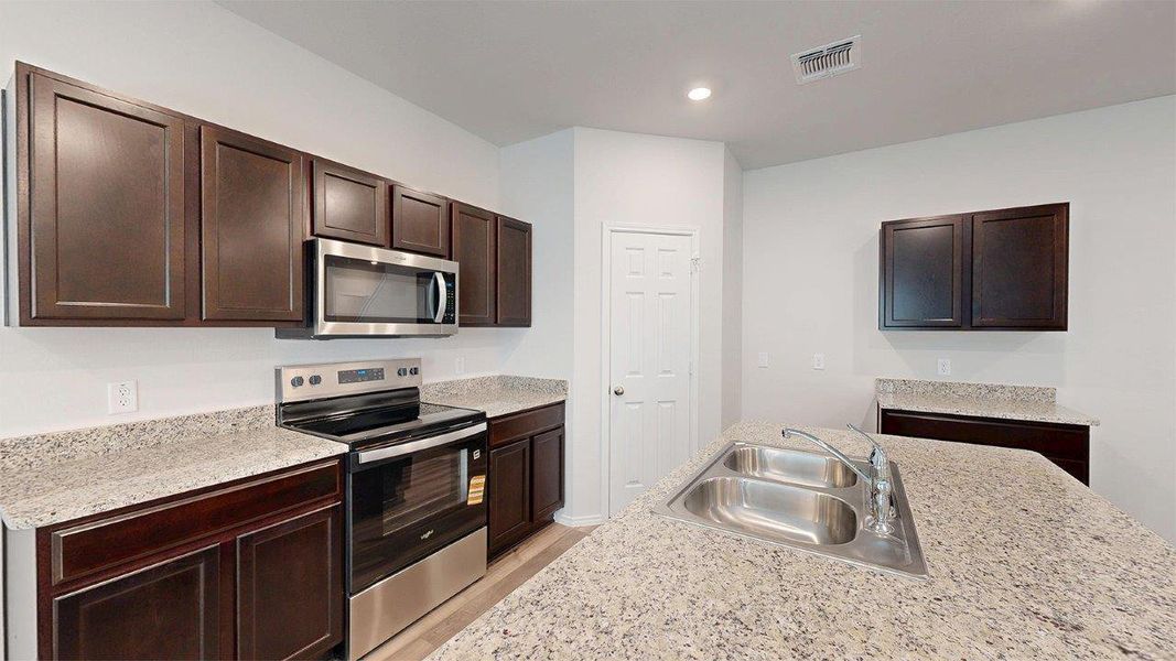 Kitchen featuring an island with a double basin stainless steel sink, stainless steel appliances, dark wood cabinetry, and light-toned countertops Kitchen featuring an island with a double basin stainless steel sink, stainless steel appliances, dark wood cabinetry, and light-toned countertops