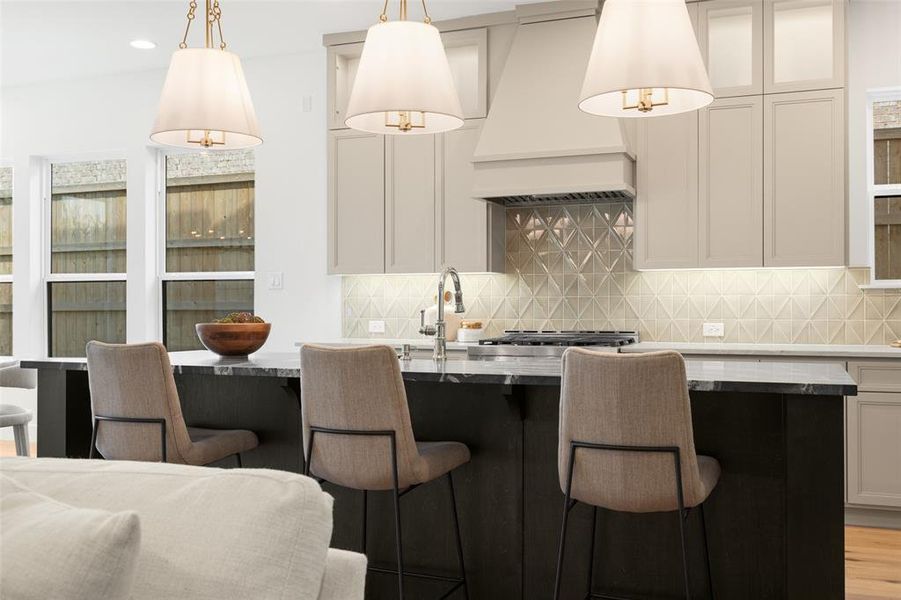 Kitchen featuring custom exhaust hood, a kitchen bar, backsplash, and hanging light fixtures Kitchen featuring custom exhaust hood, a kitchen bar, backsplash, and hanging light fixtures