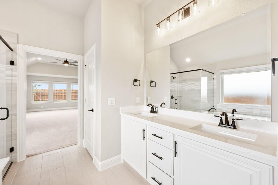 Ensuite bathroom with double vanity, white cabinets, glass shower, and view into carpeted bedroom Ensuite bathroom with double vanity, white cabinets, glass shower, and view into carpeted bedroom