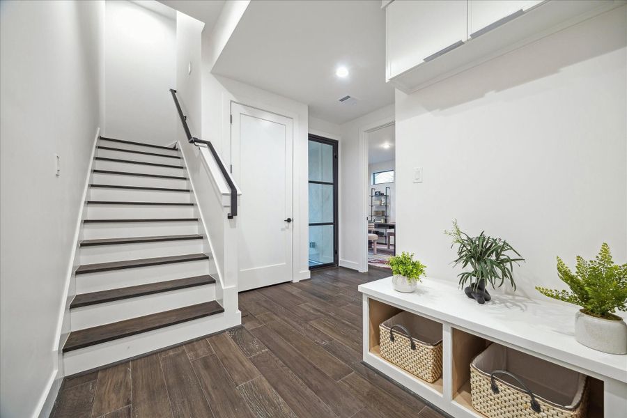 Welcoming first-floor entry featuring clean architectural lines, modern white walls, and rich wood-look tile flooring that flows seamlessly throughout. A sleek staircase with contrasting dark handrail leads to the main living level, while a glass-paneled door allows natural light to filter in. Thoughtfully designed built-in bench seating with storage cubbies adds both style and functionality to this bright, contemporary foyer.