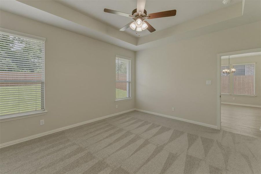Spacious room featuring a tray ceiling, ceiling fan with integrated lighting, two windows with blinds, light tan wall paint, and patterned carpet flooring