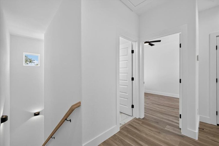 Hallway featuring wood-finish flooring, white paneled doors with black hardware, and a staircase with a natural wood handrail Hallway featuring wood-finish flooring, white paneled doors with black hardware, and a staircase with a natural wood handrail