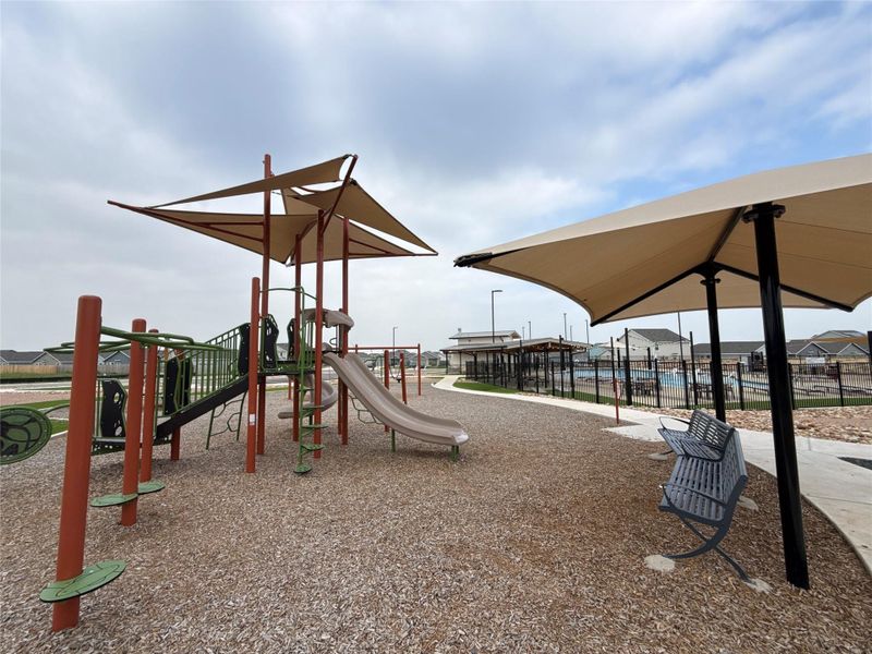 Community playground featuring modern play equipment, shade canopies, wood chip surfacing, and a nearby pool area