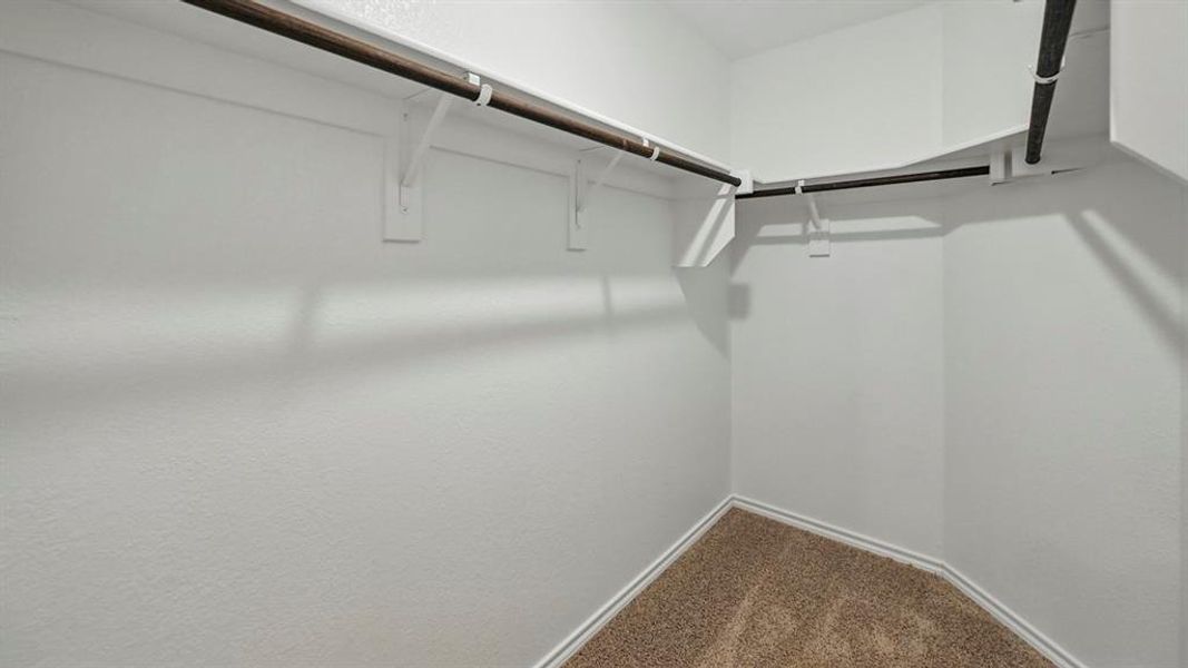 Walk-in closet featuring light-colored textured walls, multiple metal clothing rods, and neutral-toned carpet flooring