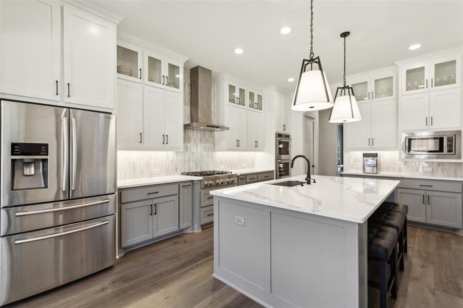 Kitchen with wall chimney range hood, a kitchen island with sink, stainless steel appliances, gray cabinetry, and decorative light fixtures