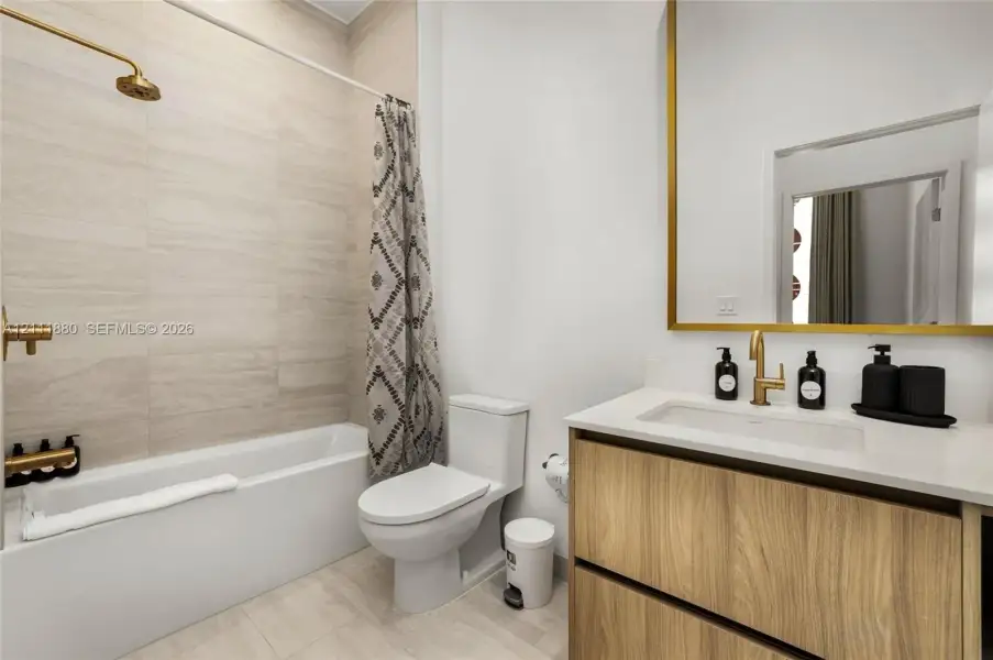 Bathroom featuring a wood-finish vanity with a stone countertop, a large rectangular mirror with a gold-toned frame, wall-mounted gold-toned plumbing fixtures, and floor-to-ceiling tile surrounding the bathtub