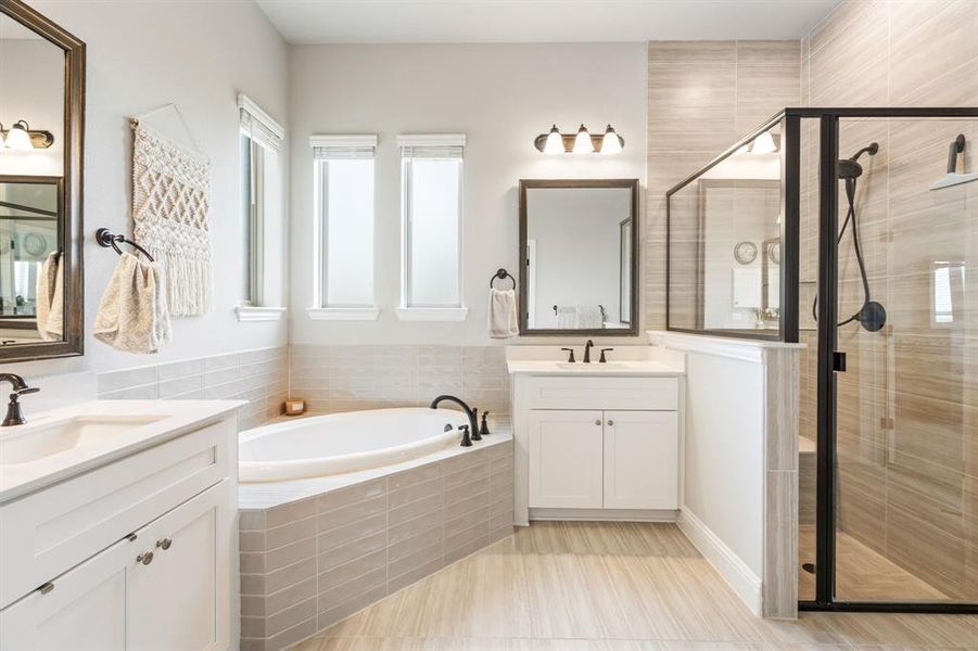 Bathroom featuring a corner soaking tub with tile surround, a walk-in shower with clear glass enclosure and dark trim, two distinct vanity areas with integrated sinks and white cabinetry, and light-toned tile flooring Bathroom featuring a corner soaking tub with tile surround, a walk-in shower with clear glass enclosure and dark trim, two distinct vanity areas with integrated sinks and white cabinetry, and light-toned tile flooring
