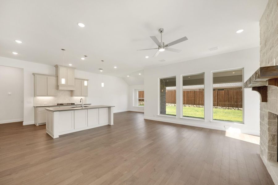 Open-concept living area with hardwood floors, kitchen island, stone fireplace, and ceiling fan