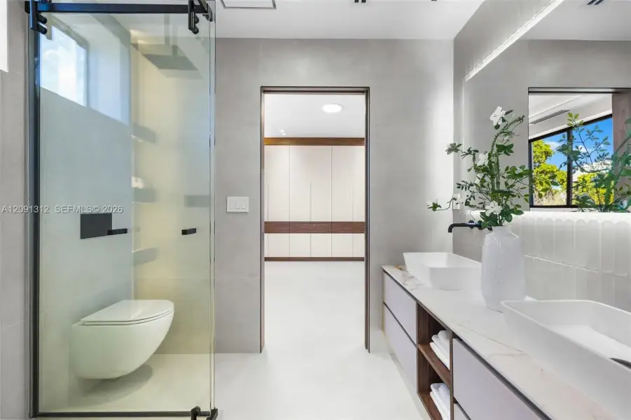 Bathroom featuring a glass-enclosed shower with matte black hardware, wall-mounted commode, and dual-sink vanity with light-toned countertop and open shelving Bathroom featuring a glass-enclosed shower with matte black hardware, wall-mounted commode, and dual-sink vanity with light-toned countertop and open shelving