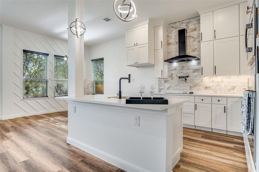 Kitchen featuring tasteful backsplash, white cabinets, a center island with sink, wall chimney exhaust hood, and light wood-style flooring