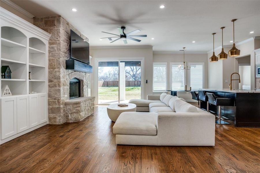 Living room with built in shelves, a stone fireplace, dark hardwood / wood-style floors, crown molding, and ceiling fan with notable chandelier