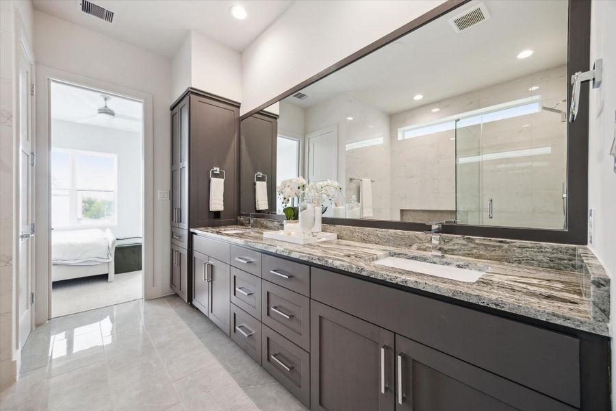 This modern bathroom features a spacious double vanity with elegant marble countertops and ample storage. A large mirror enhances the space, and the room is well-lit with recessed lighting. The adjoining bedroom is visible through an open door, providing a seamless connection between the spaces.