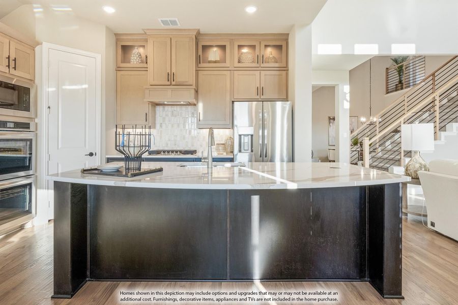 Kitchen with large dark island, white countertops, tan cabinets, stainless appliances, and staircase visible in background Kitchen with large dark island, white countertops, tan cabinets, stainless appliances, and staircase visible in background