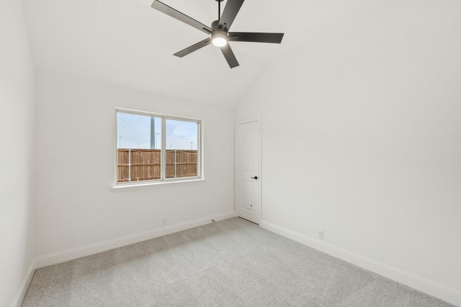 Empty bedroom with gray carpet, ceiling fan, white walls, and a window overlooking a wood fence