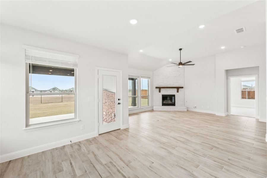 Open living room with light wood floors, brick fireplace, ceiling fan, and backyard views through large windows Open living room with light wood floors, brick fireplace, ceiling fan, and backyard views through large windows