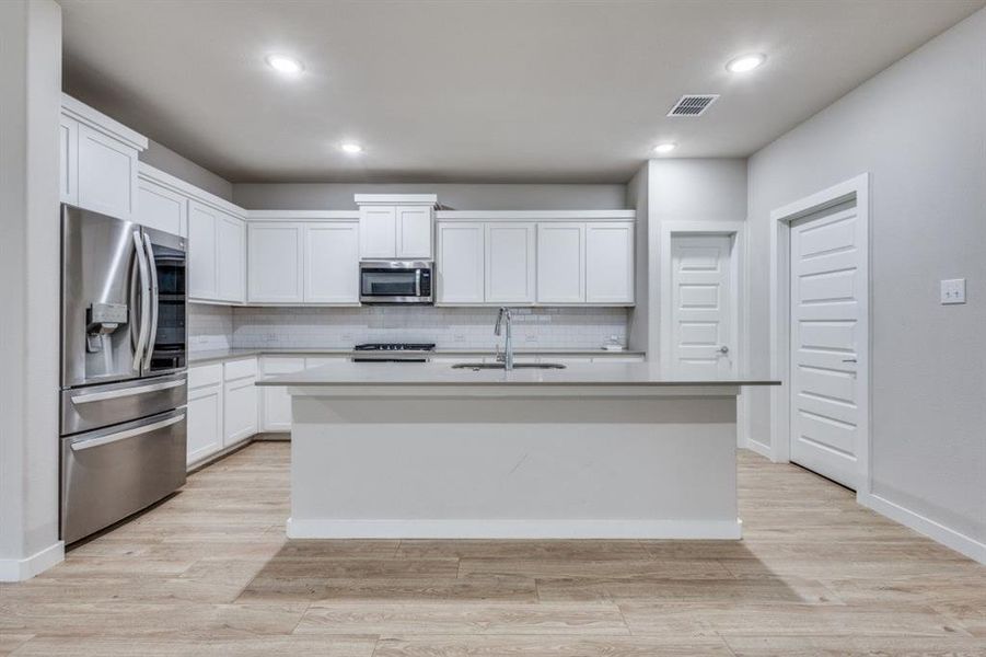 Kitchen featuring white cabinetry, appliances with stainless steel finishes, decorative backsplash, a center island with sink, and light wood-type flooring Kitchen featuring white cabinetry, appliances with stainless steel finishes, decorative backsplash, a center island with sink, and light wood-type flooring