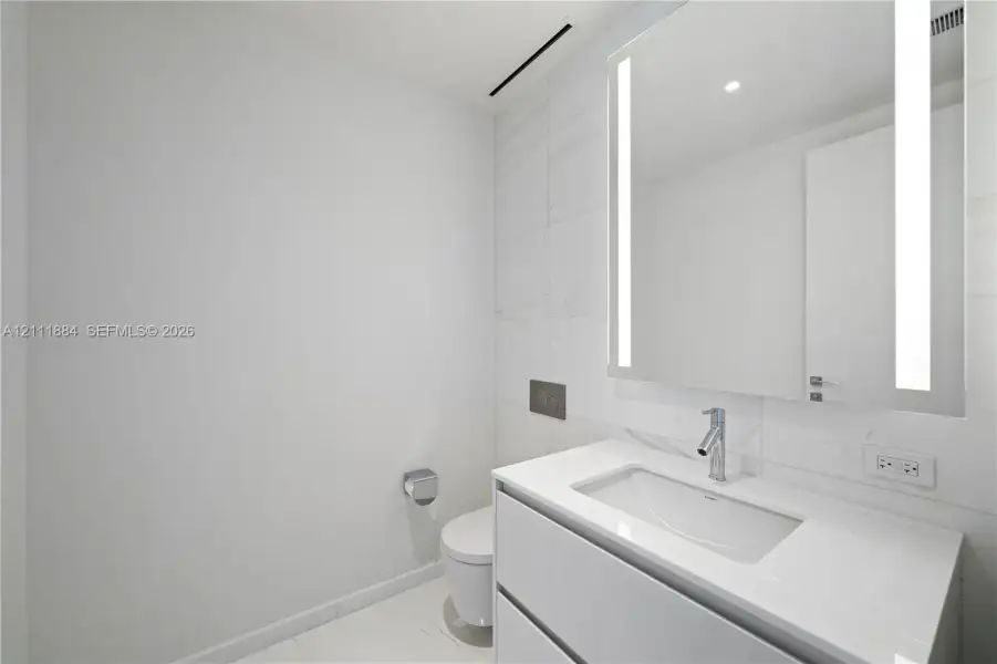 Floating vanity featuring a white countertop and rectangular basin, integrated LED-backlit mirror, wall-mounted faucet, and large-format tile wall Floating vanity featuring a white countertop and rectangular basin, integrated LED-backlit mirror, wall-mounted faucet, and large-format tile wall