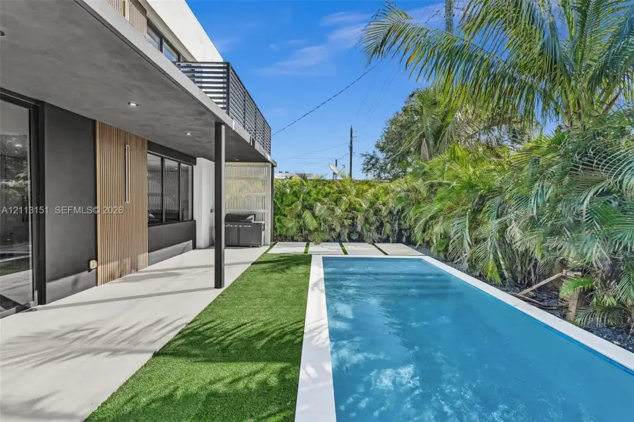 Modern exterior patio featuring concrete flooring, a recessed ceiling, wood-toned wall cladding, an artificial turf lawn, and a rectangular swimming pool with a white border Modern exterior patio featuring concrete flooring, a recessed ceiling, wood-toned wall cladding, an artificial turf lawn, and a rectangular swimming pool with a white border