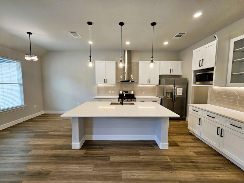 Kitchen featuring stainless steel appliances, an island with sink, decorative light fixtures, white cabinets, and wall chimney range hood