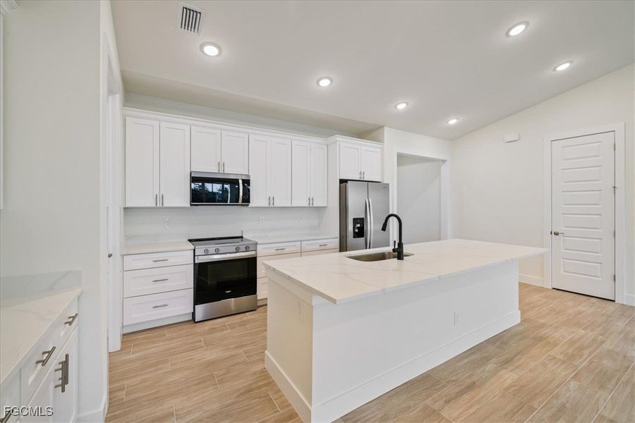 Kitchen featuring appliances with stainless steel finishes, white cabinets, wood finish floors, light stone countertops, and an island with sink