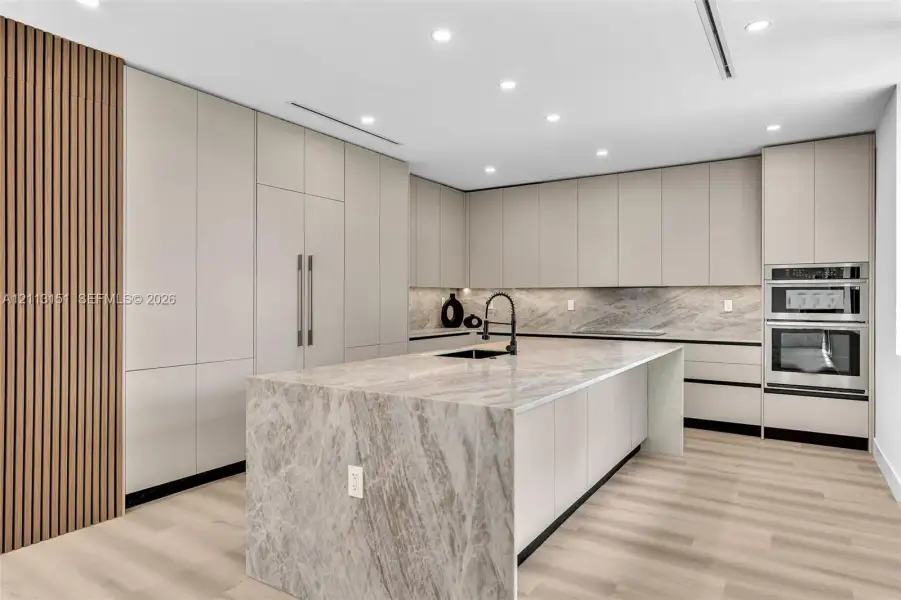 Kitchen featuring a center island with a stone waterfall edge, slab stone backsplash, flat-panel cabinetry, vertical wood slat accent wall, and built-in stainless steel double wall ovens Kitchen featuring a center island with a stone waterfall edge, slab stone backsplash, flat-panel cabinetry, vertical wood slat accent wall, and built-in stainless steel double wall ovens