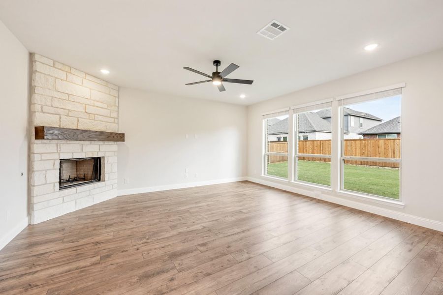 Open living room with stone fireplace, hardwood floors, ceiling fan, and large windows overlooking backyard