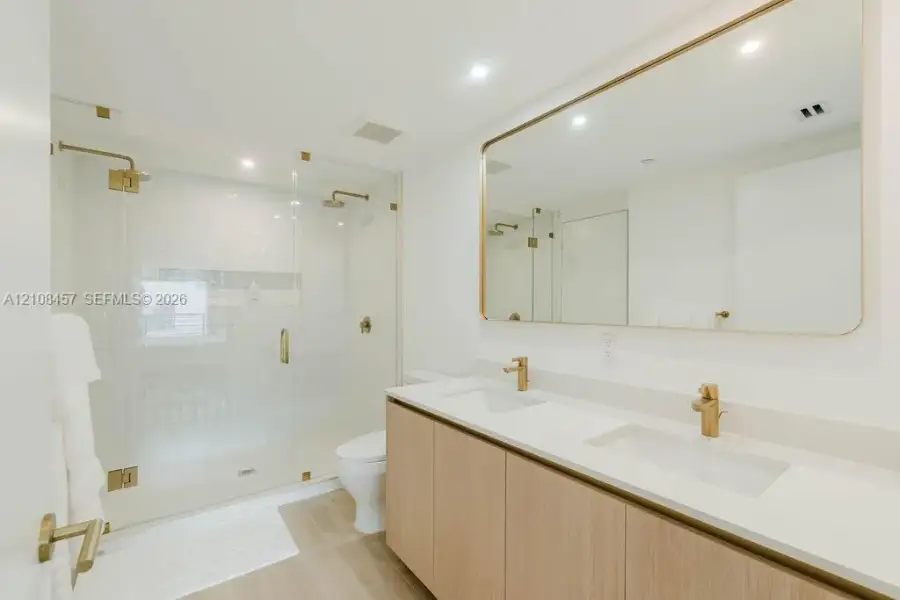 Double vanity with light wood-finish cabinetry, quartz countertop, dual undermount sinks, brass fixtures, and a glass-enclosed shower Double vanity with light wood-finish cabinetry, quartz countertop, dual undermount sinks, brass fixtures, and a glass-enclosed shower