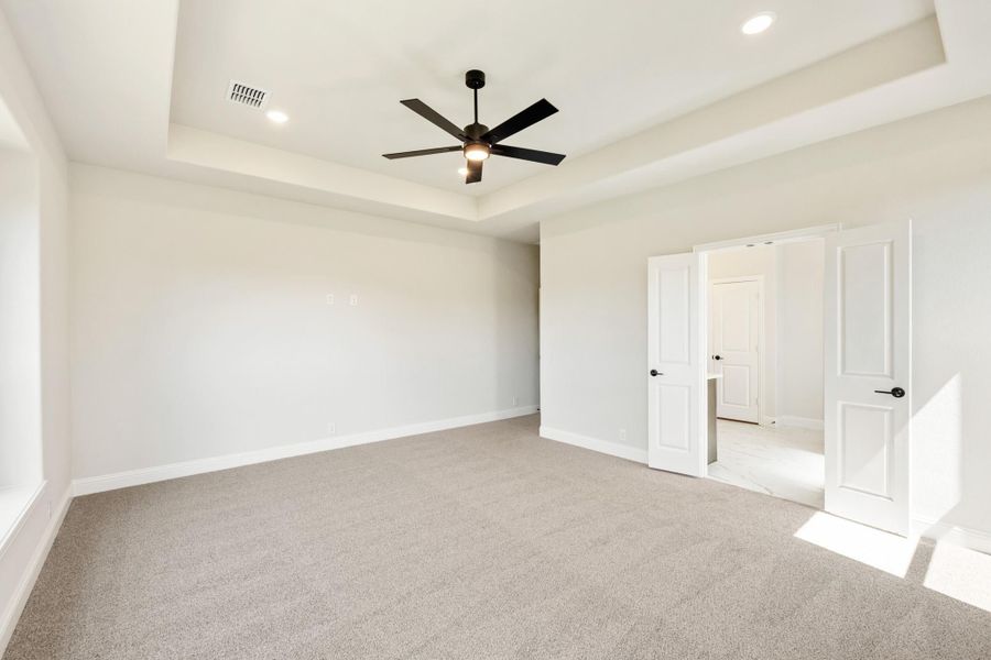 Empty bedroom with tray ceiling, black ceiling fan, carpet flooring, and double doors leading to bathroom