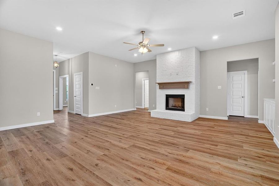 Spacious interior room featuring wood-finish flooring, a white brick fireplace with a wood mantel, recessed lighting, and a ceiling fan Spacious interior room featuring wood-finish flooring, a white brick fireplace with a wood mantel, recessed lighting, and a ceiling fan
