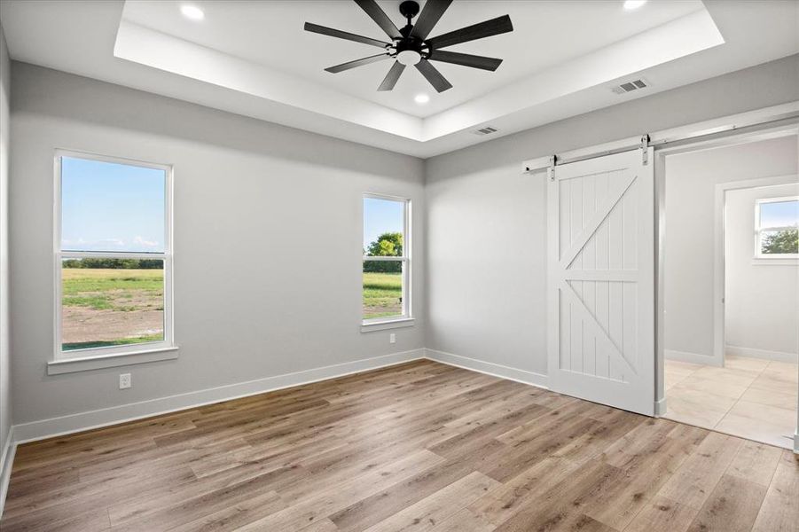 Unfurnished bedroom with a raised ceiling, light wood finished floors, a barn door, and recessed lighting Unfurnished bedroom with a raised ceiling, light wood finished floors, a barn door, and recessed lighting