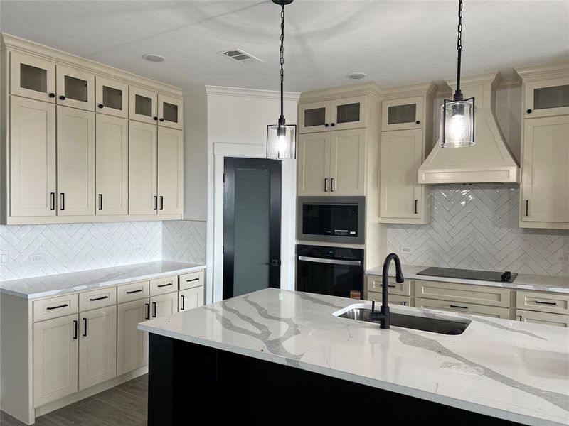 Kitchen featuring custom range hood, light stone counters, cream cabinets, black appliances, and pendant lighting