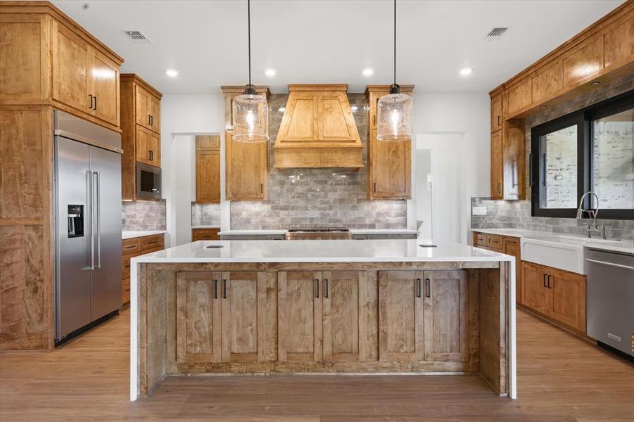 Kitchen featuring a large island, light stone countertops, brown cabinets, built in appliances, and backsplash