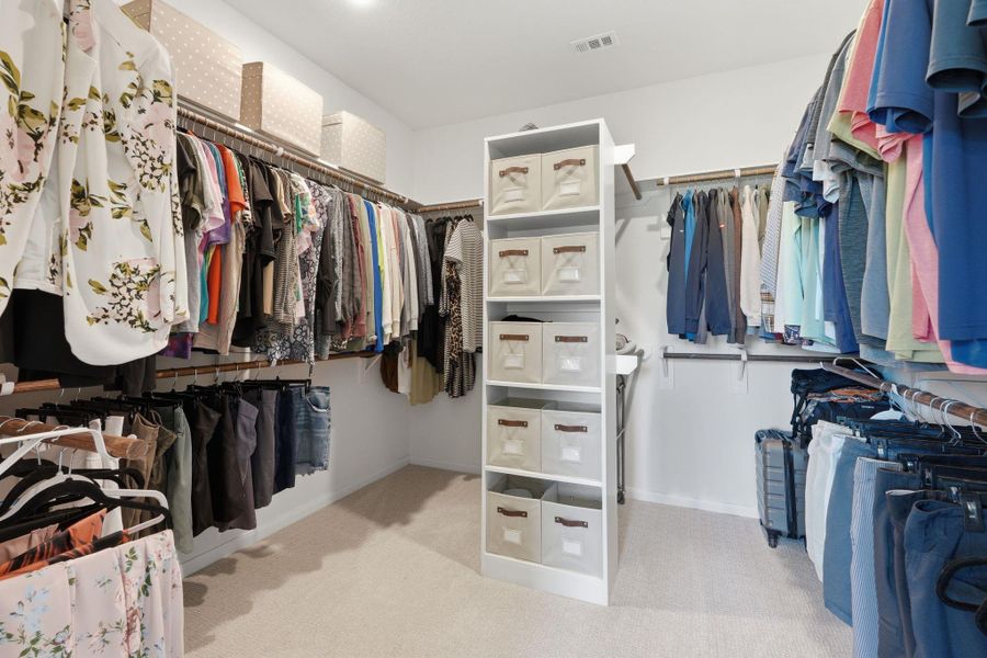 This photo showcases a spacious, well-organized walk-in closet with ample hanging space, shelving units, and storage bins for efficient organization. The room is well-lit and carpeted, providing a clean and accessible storage solution for a variety of clothing and accessories.