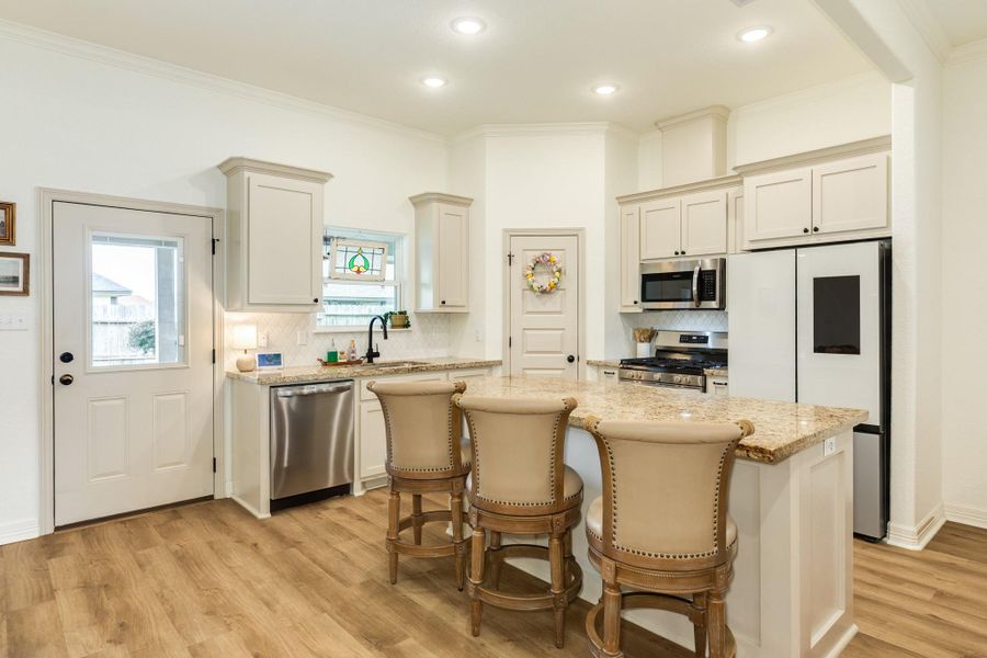 This kitchen features a bright, open layout with modern appliances, granite countertops, and a cozy breakfast bar with seating. Light wood look flooring complements the neutral cabinetry, and a small stained glass window adds charm. Ideal for both cooking and entertaining.