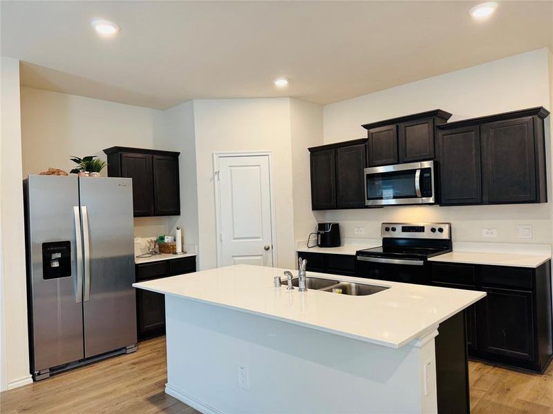 Kitchen with appliances with stainless steel finishes, a kitchen island with sink, light wood-type flooring, dark cabinetry, and recessed lighting Kitchen with appliances with stainless steel finishes, a kitchen island with sink, light wood-type flooring, dark cabinetry, and recessed lighting