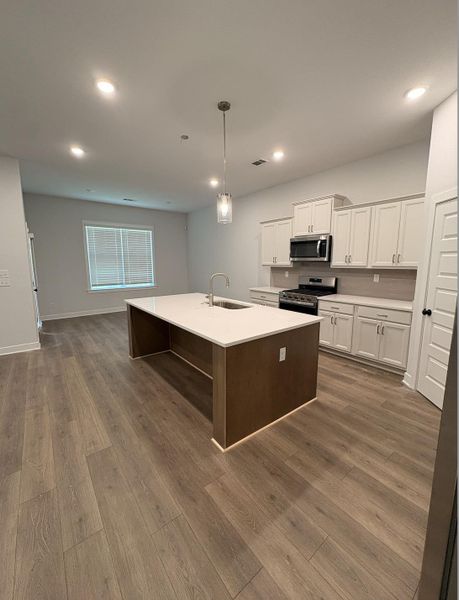 Expansive kitchen island with a white countertop and undermount sink, stainless steel appliances, white cabinetry, wood-finish flooring, and recessed lighting