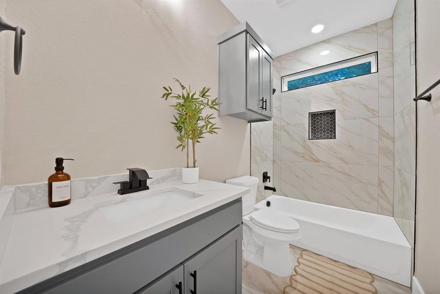 Bathroom featuring a vanity with a white countertop and dark gray cabinetry, a matte black faucet, a white bathtub, and light-toned tiled walls with a decorative accent niche Bathroom featuring a vanity with a white countertop and dark gray cabinetry, a matte black faucet, a white bathtub, and light-toned tiled walls with a decorative accent niche