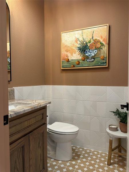Powder room featuring a wood-finish vanity with a stone countertop, a white toilet, geometric wall tiling, and patterned floor tiling