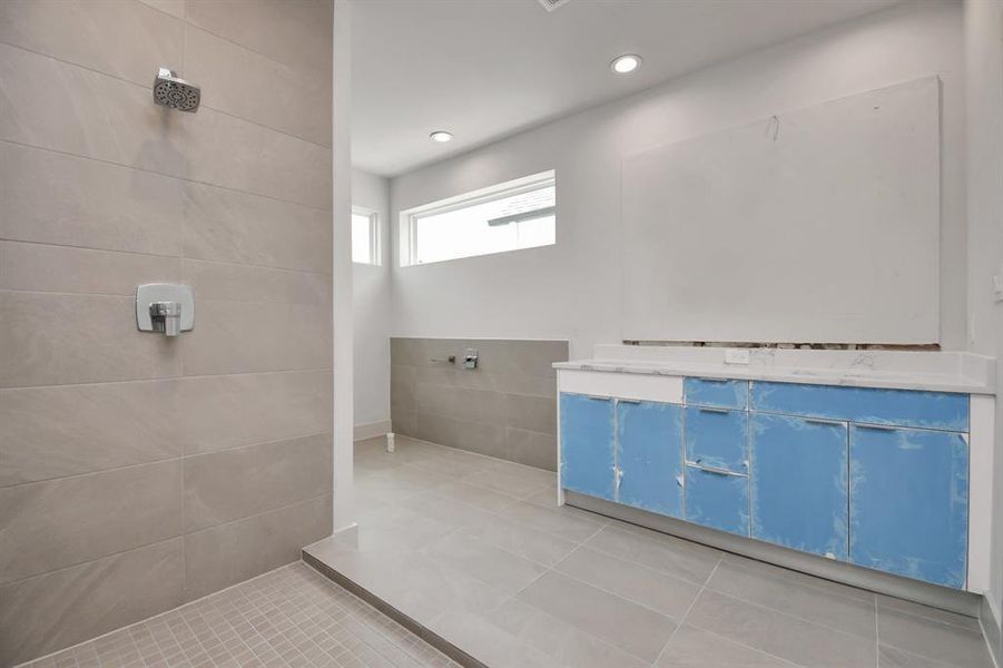 Spacious and modern master bathroom under with a large walk-in shower and tile throughout, recessed lighting, and a dual vanity sink. A high window allows for natural light while maintaining privacy.