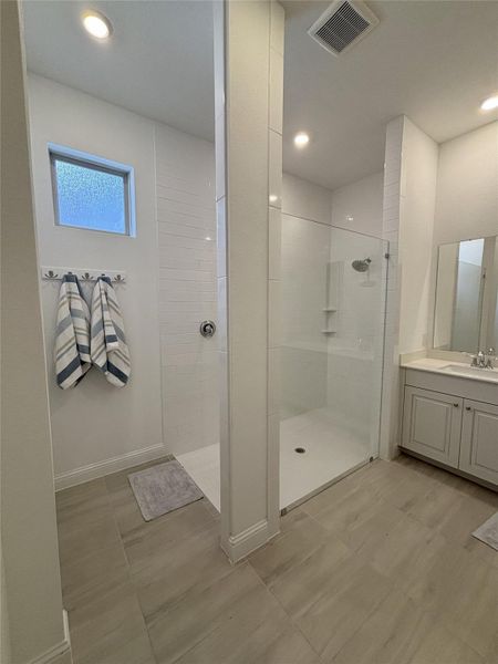 This bathroom features a modern, spacious design with a large walk-in shower enclosed in glass. The space is bright with recessed lighting and a frosted window for privacy. It includes a sleek vanity with ample counter space. The neutral color palette and tiled flooring create a clean and inviting atmosphere.
