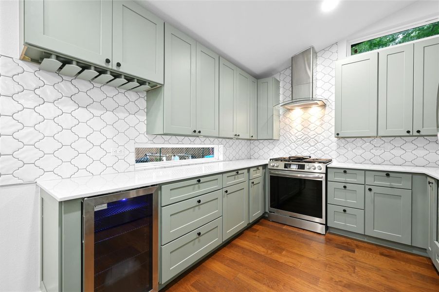 Kitchen with stainless steel gas range oven, beverage cooler, wall chimney exhaust hood, light countertops, and vaulted ceiling