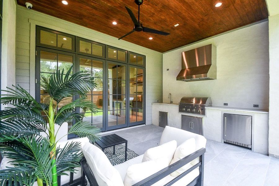 This photo showcases a modern outdoor patio with a built-in stainless steel grill, sink, and mini-fridge. It features comfortable seating, a ceiling fan, and large glass doors that open to the interior, making it perfect for entertaining.