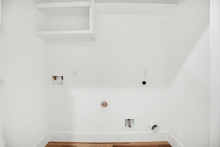 This photo shows a laundry area with hookups for a washer and dryer. It features white walls, an overhead shelf, and wood flooring.