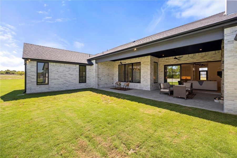 Rear view of house with ceiling fan, a patio, brick siding, a shingled roof, and a lawn