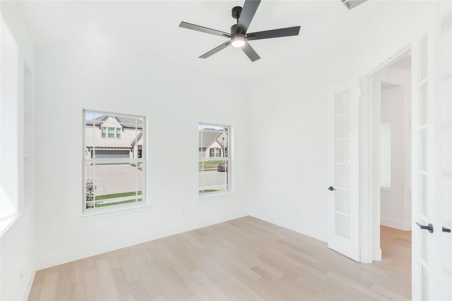 Empty room with white walls, light hardwood floors, ceiling fan, and two windows with neighborhood view